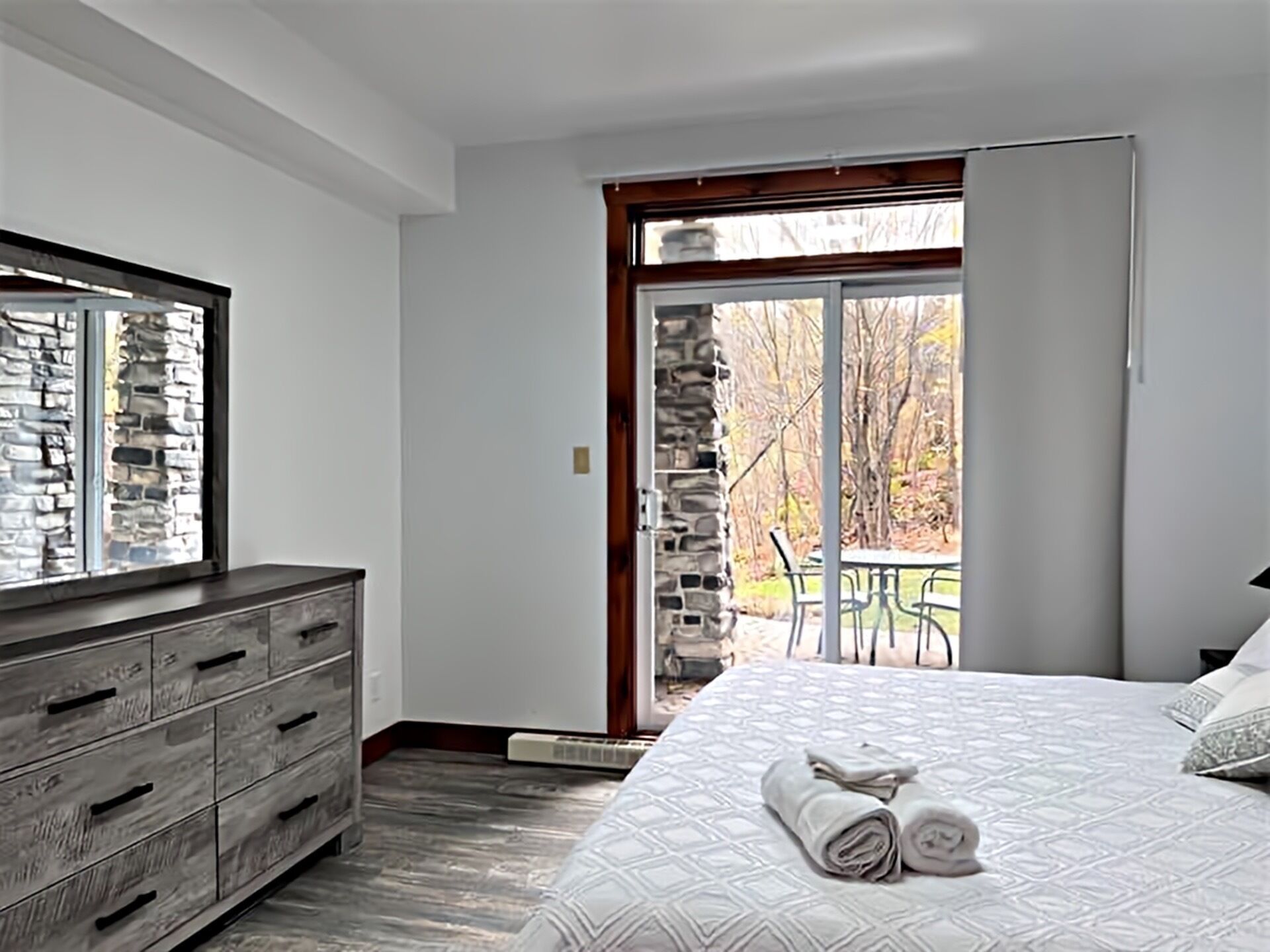 A bedroom with a large bed, a dresser, a stone wall, and a view of the outdoors.