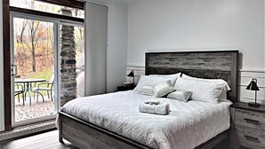 Un dormitorio con una cama grande, una pared de piedra, un ventanal corredero a un patio y una mesita de noche con una lámpara.