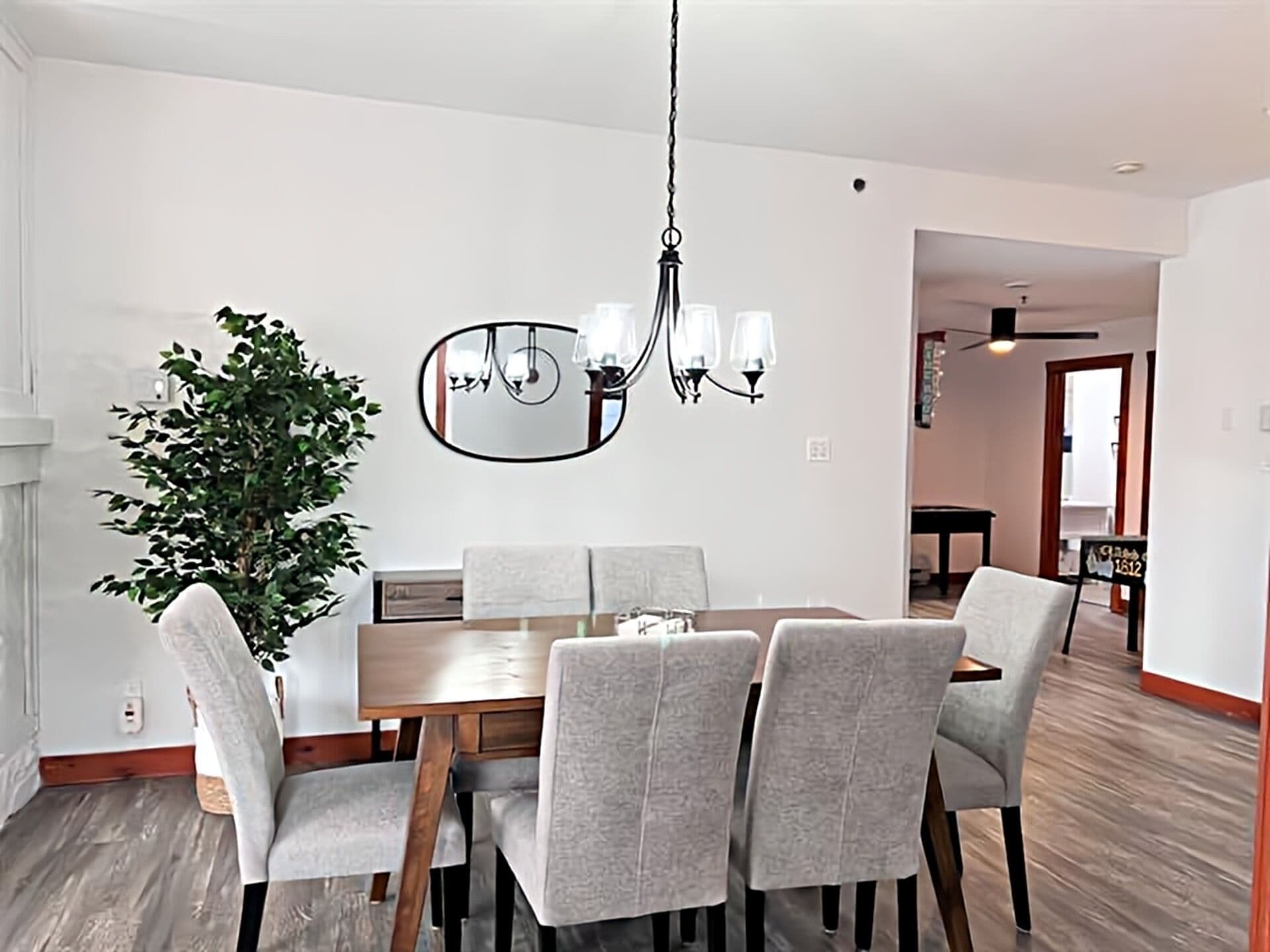 A dining area with a wooden table, grey upholstered chairs, a hanging chandelier, a large mirror, and a potted plant.
