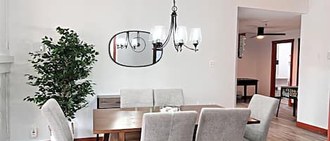 Comedor con mesa de madera, sillas tapizadas en gris, candelabro colgante, espejo grande y planta en maceta.