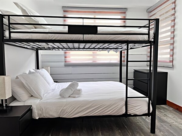 A bunk bed with white bedding, a bedside table with a lamp, and a window with blinds. - Les Eaux 227-1 by Escapades Tremblant (Mont-Tremblant)