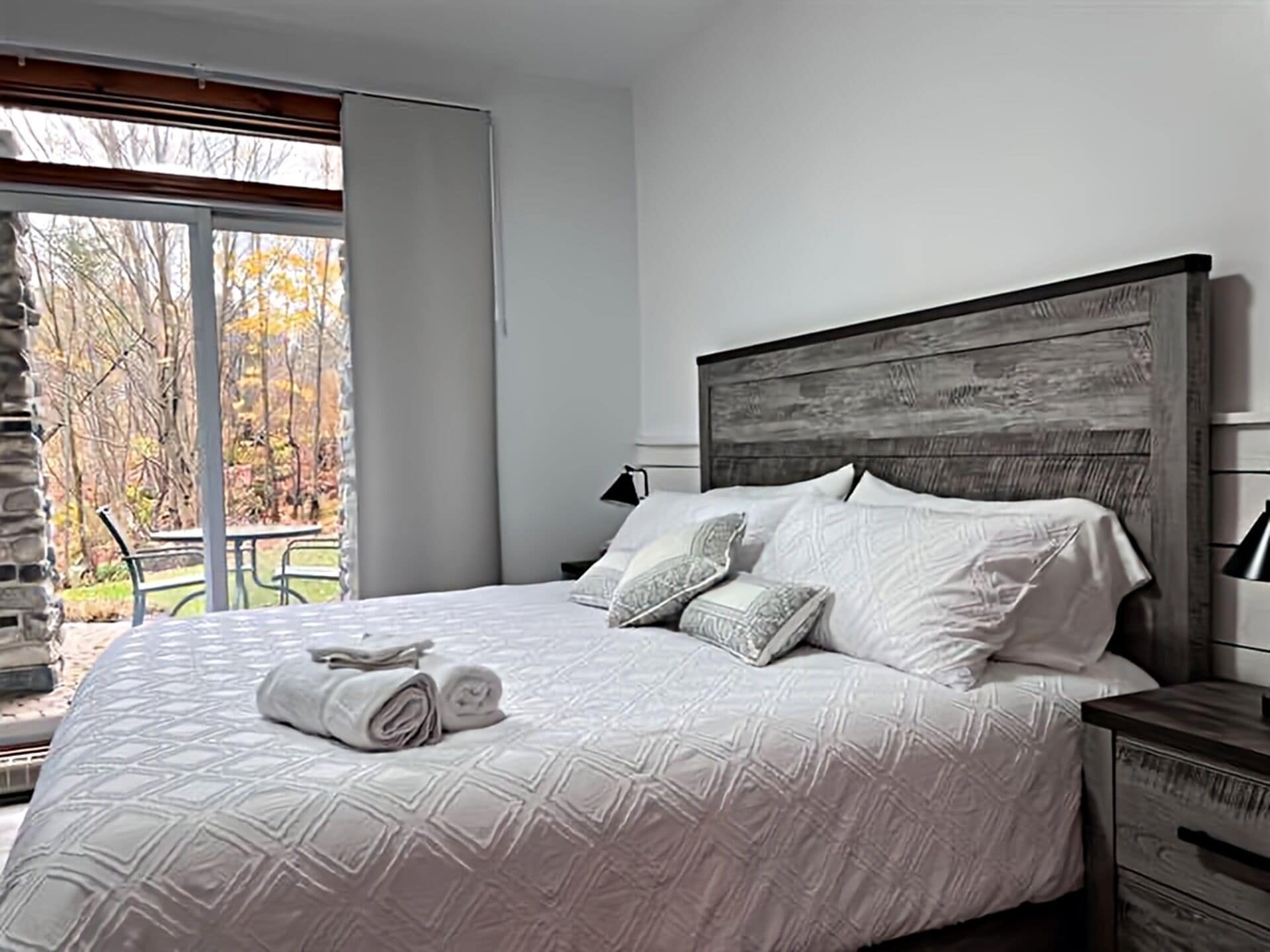 A bedroom with a large bed, a wooden headboard, a nightstand, and a view of a patio with a table and chairs.