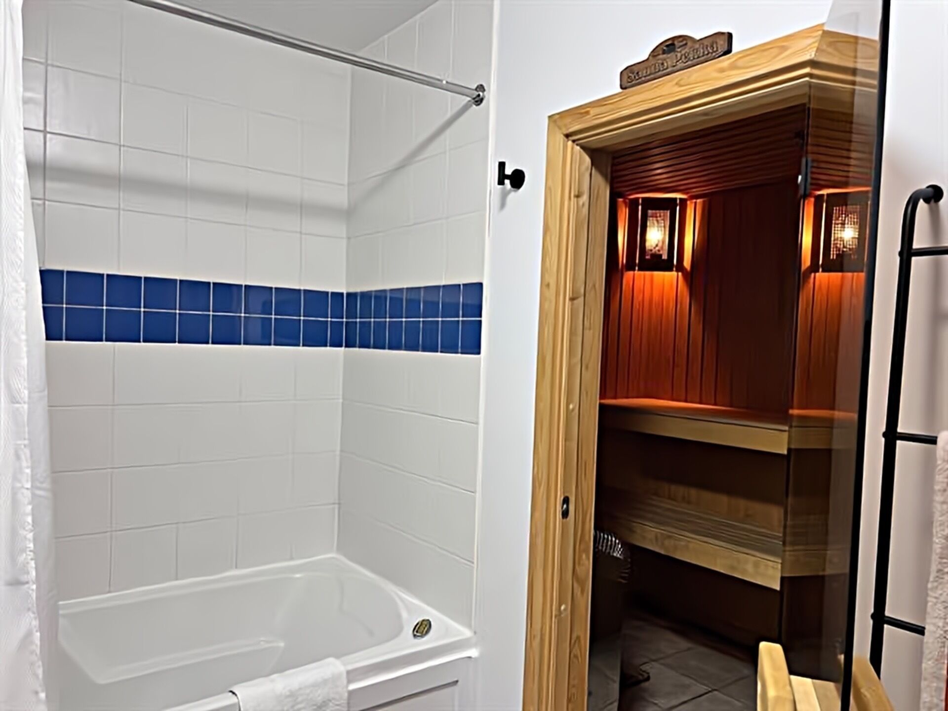 Et badeværelse med et hvidt badekar, et bruseområde med blå fliser og et trækommode med et spejl.