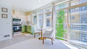 A modern kitchen with white cabinets, a dining area with a white table and chairs, and large windows with a view of greenery.