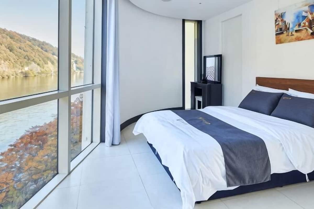 A bedroom with a large window, a bed, a TV, and a painting on the wall.