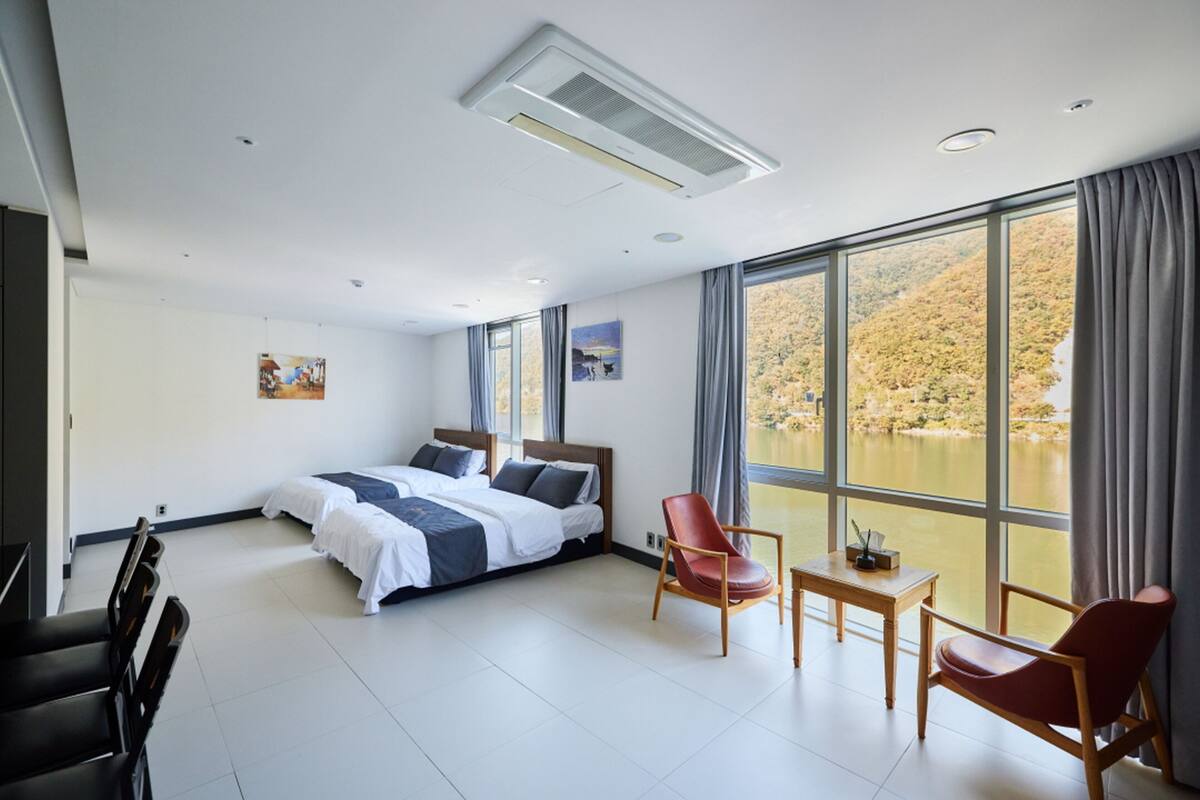 A modern hotel room with a large bed, two chairs, a small table, and a view of a lake through large windows.