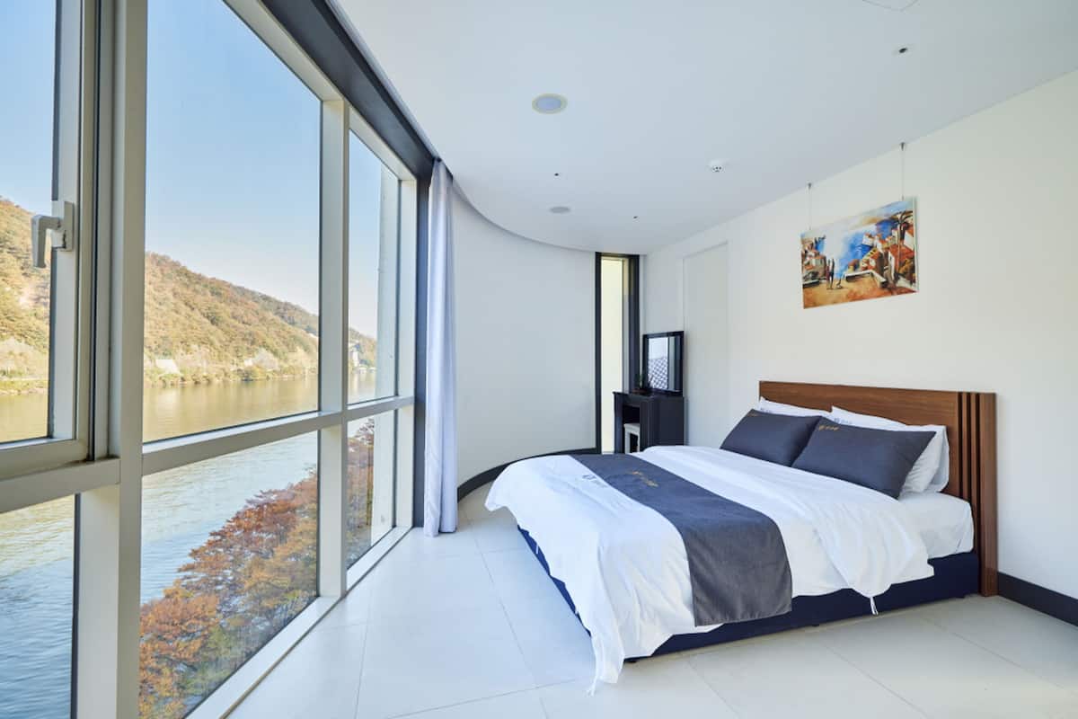 A modern bedroom with a large window, a bed, and a painting on the wall.