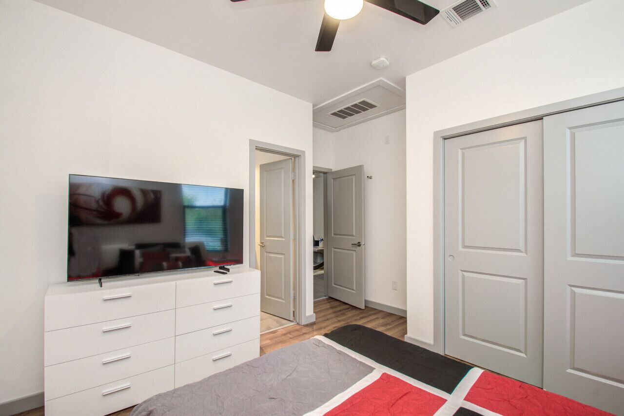 A modern room with a flat-screen TV on a white dresser, a ceiling fan, and a hallway leading to a door.