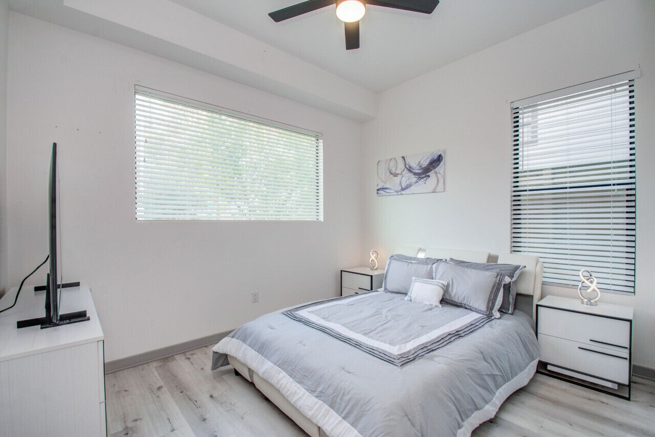 A bedroom with a bed, two bedside tables, a ceiling fan, and two windows with blinds.