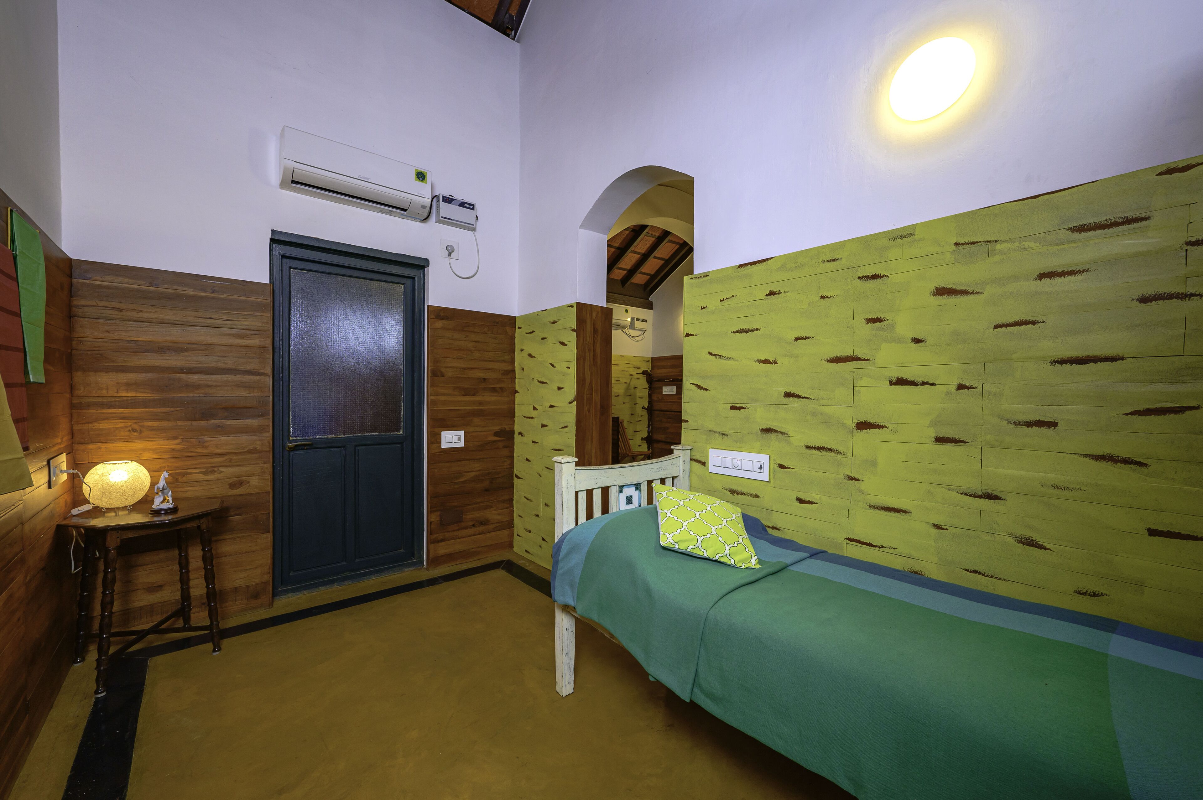 A room with a bed, a wooden door, an air conditioning unit, and a lamp on a small table.