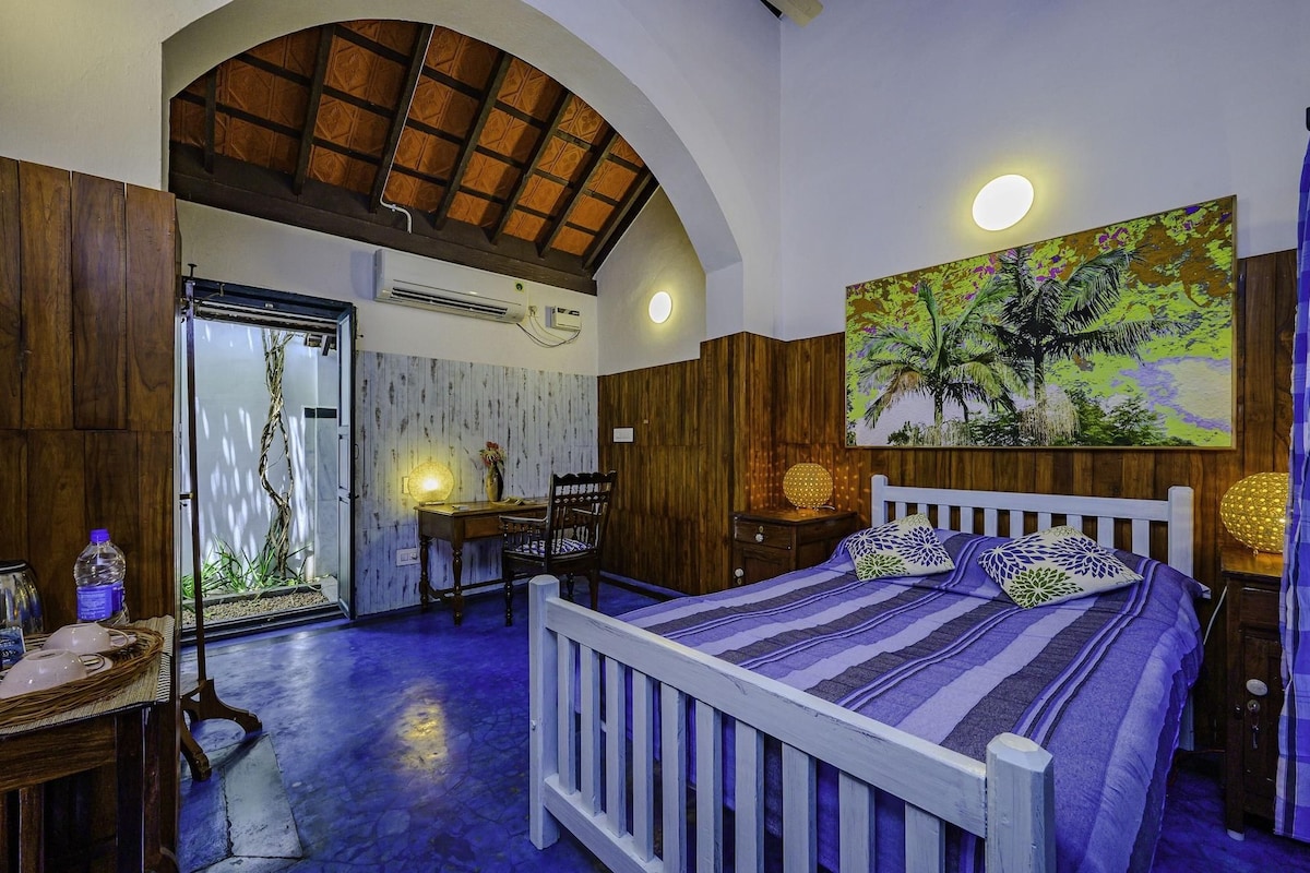 A bedroom with a wooden ceiling, a bed with patterned bedding, a desk with a lamp, and a painting on the wall.
