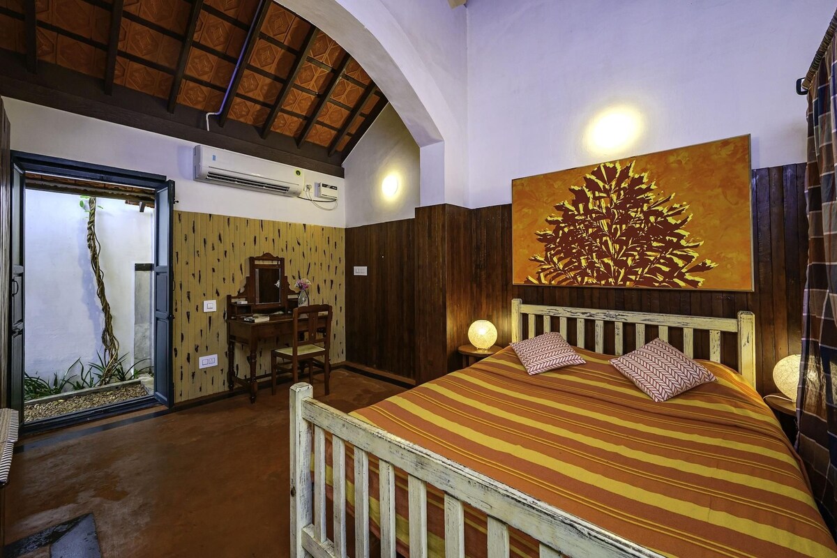 A room with a bed, a desk, and a chair. There is a painting on the wall and an air conditioning unit on the ceiling.