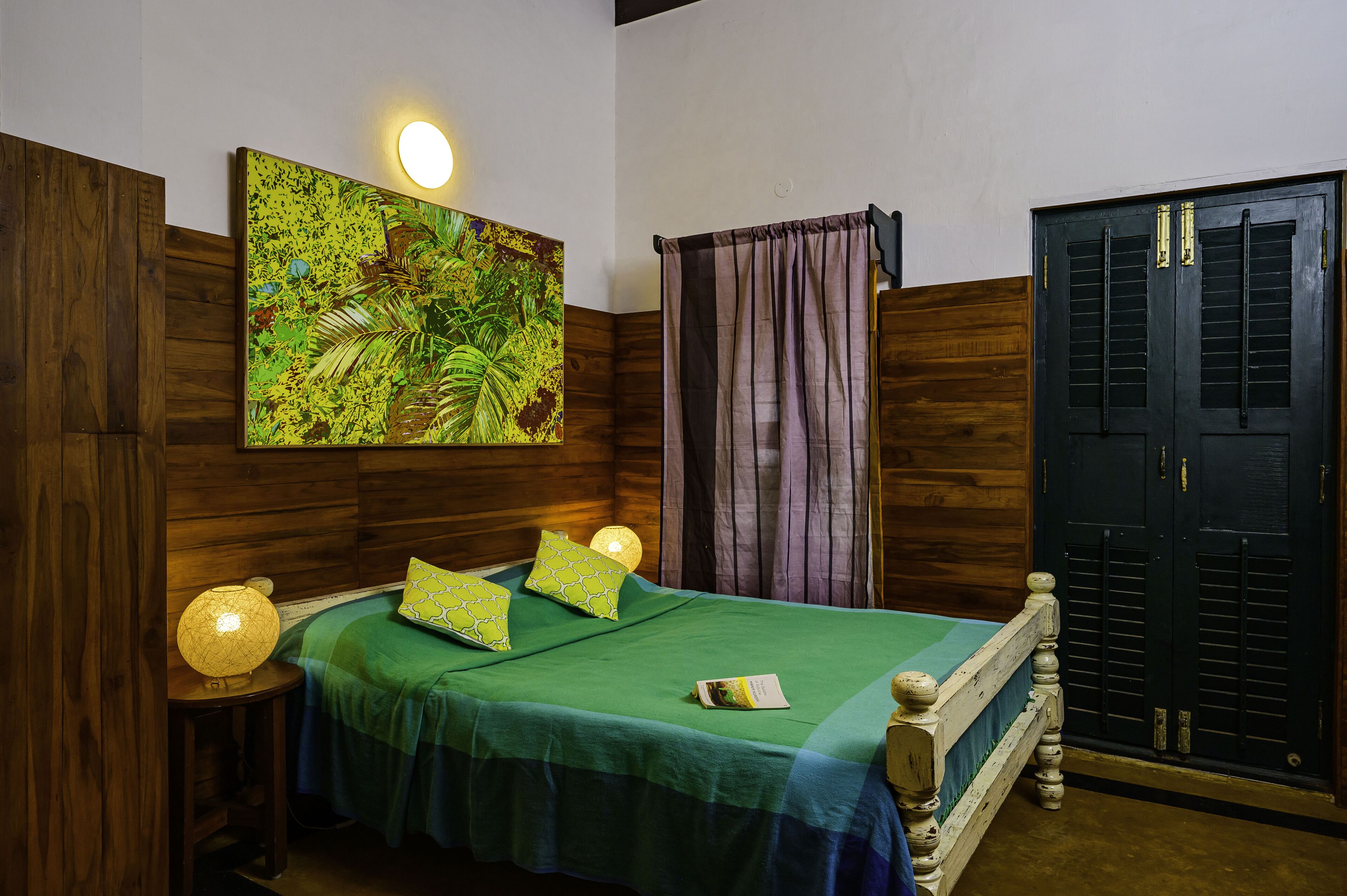 A room with a bed, a wooden headboard, a green bedspread, two pillows, a wall-mounted light, a door, and a curtain.