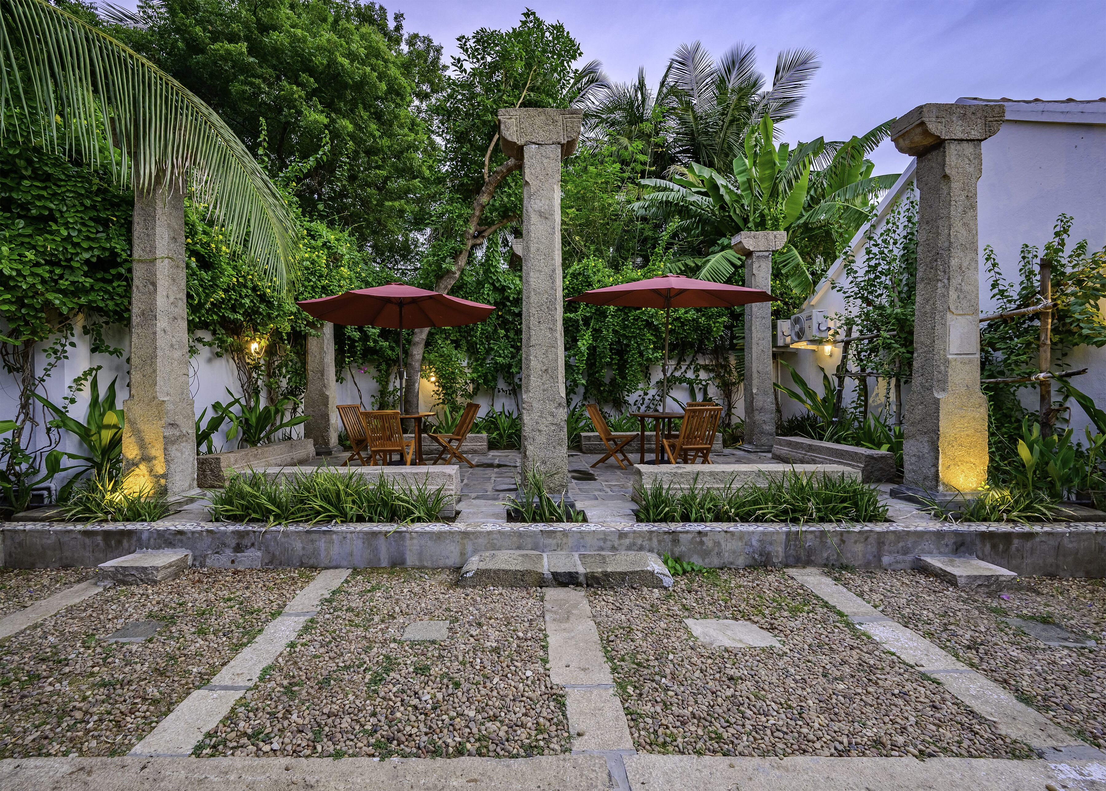 A courtyard with stone columns, a table with chairs, and a red umbrella.