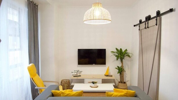 Ett modernt vardagsrum med en grå soffa, gula kuddar, ett vitt soffbord, en platt-TV, en hängande lampa och en krukväxt.