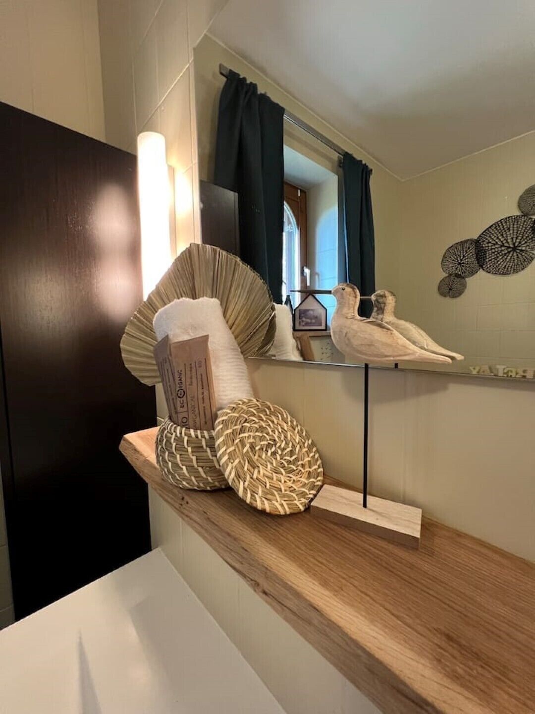 A bathroom with a wooden shelf holding decorative items, a mirror, and a window with curtains.