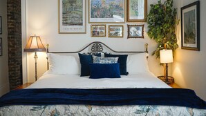 A neatly made bed with white and blue bedding, a metal headboard, and a dark blue throw blanket. There are framed artworks on the wall above the bed, and a potted plant to the right. - Vintage Frenchmen Hideaway,6Blocks to Quarter+Jazz (New Orleans)