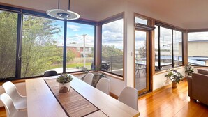 Un comedor moderno con una mesa de madera, sillas blancas y ventanas amplias con vistas a una zona residencial y un puente.