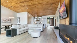 Ein modernes Wohnzimmer mit Holzbalkendecke, einem großzügigen Ecksofa, einem Kamin und einem Essbereich mit Tisch und Stühlen.