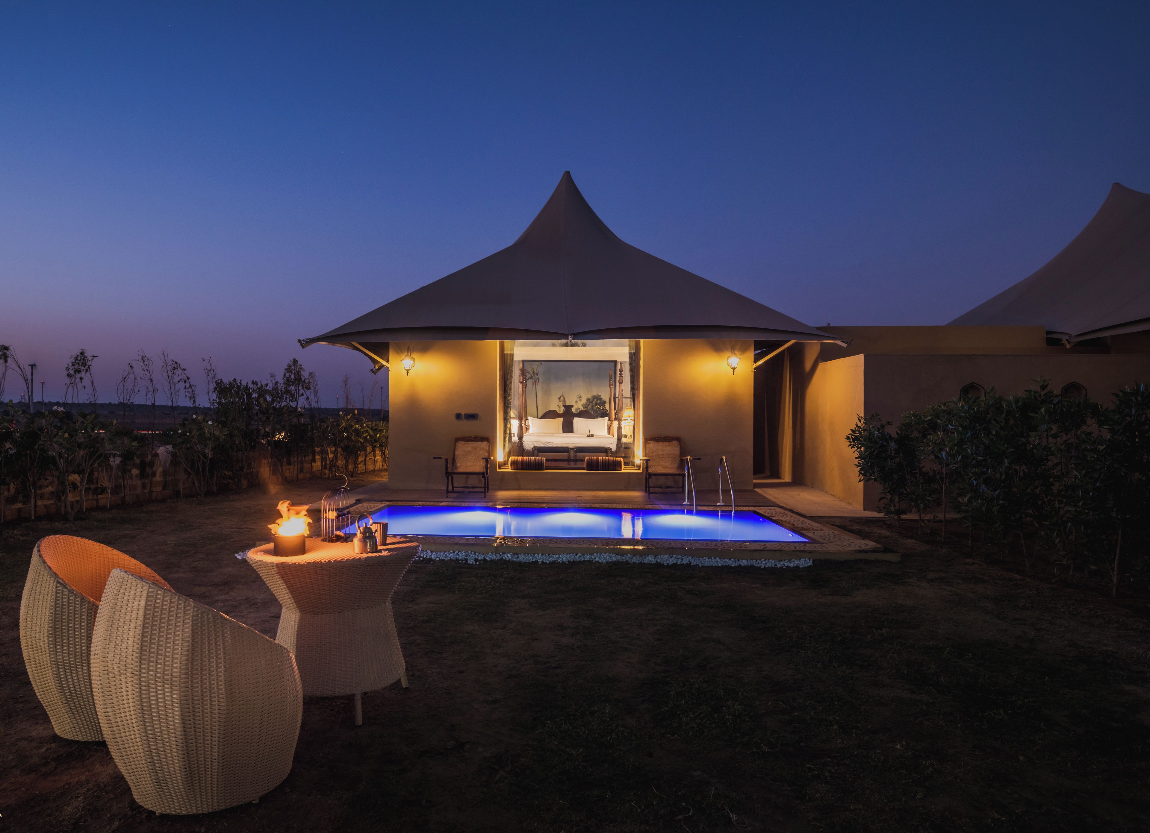 A modern outdoor pool area with a lounge chair and a table, a covered seating area, and a view of a bedroom with a large window.