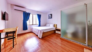 A bedroom with a wooden floor, a bed with white bedding, a desk with a chair, a TV mounted on the wall, and a window with blue curtains. - HANZ CK Saigon Central Hotel (Ho Chi Minh City)
