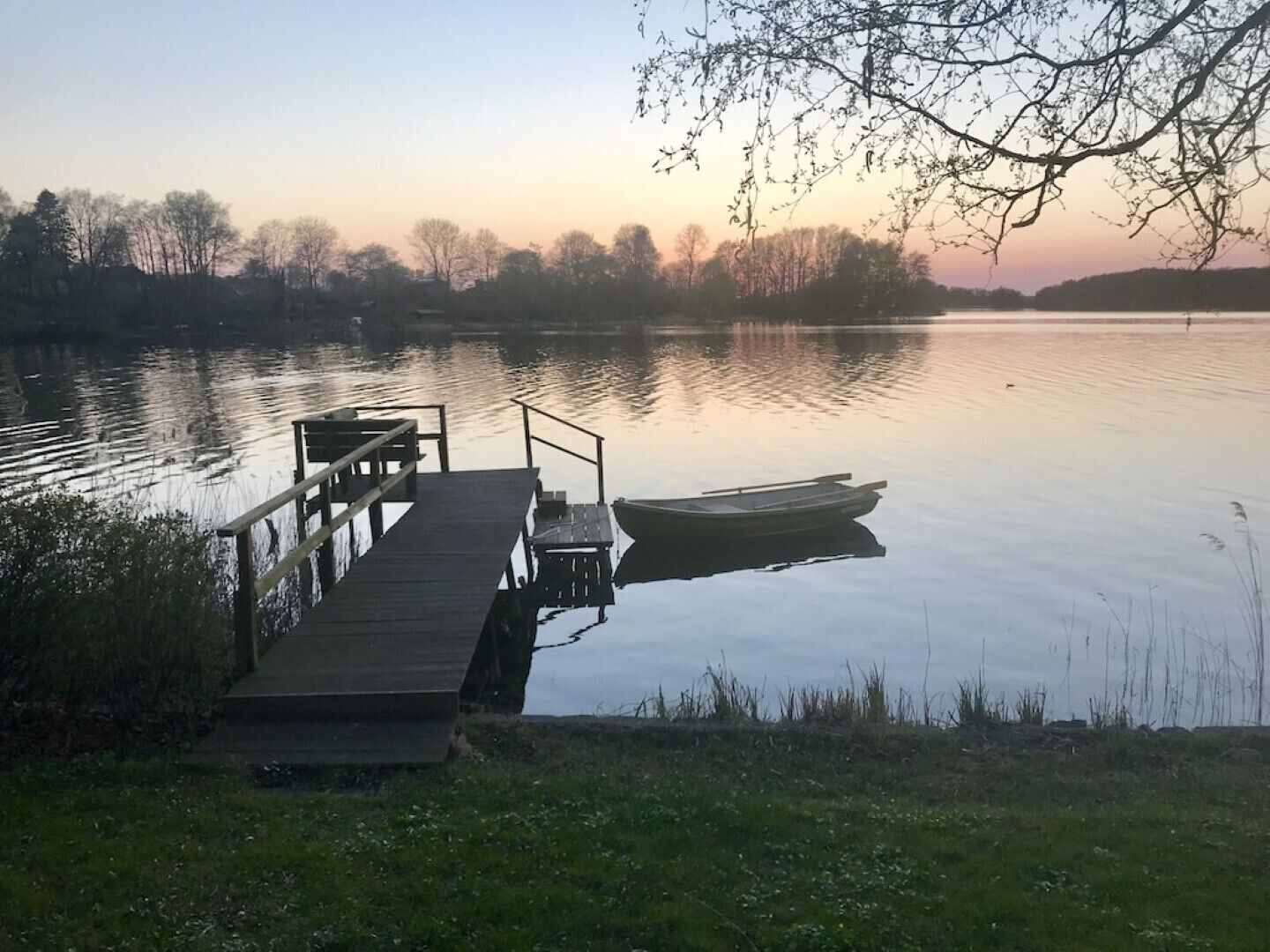 Un lac paisible, avec une jetée en bois qui s’avance sur l’eau, un petit bateau amarré à son extrémité et une branche d’arbre qui surplombe la jetée.
