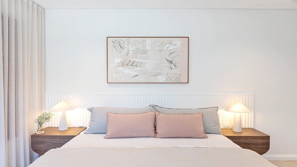 A bedroom with a bed, two bedside tables with lamps, a large abstract painting on the wall, and sheer curtains on the window.