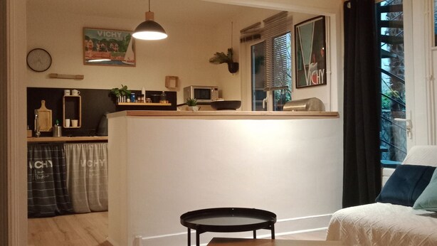 Un salon chaleureux avec une table en bois, un canapé et un coin cuisine à l’arrière-plan.