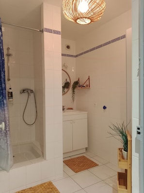 Un baño con ducha, lavamanos y espejo. Hay un perchero y un estante con artículos de aseo.