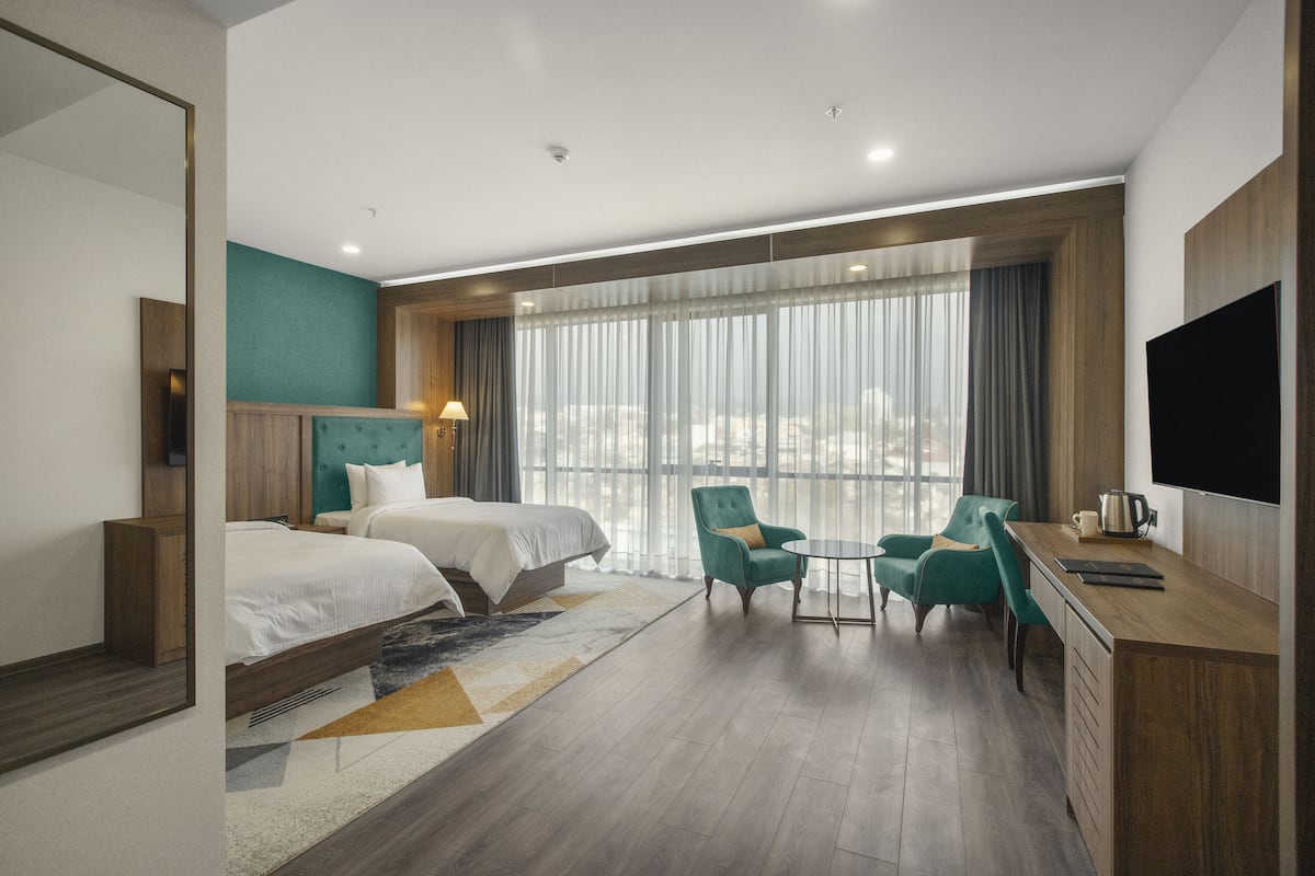 A hotel room with two beds, a desk with a flat-screen TV, a coffee maker, and a view of the city through large windows.