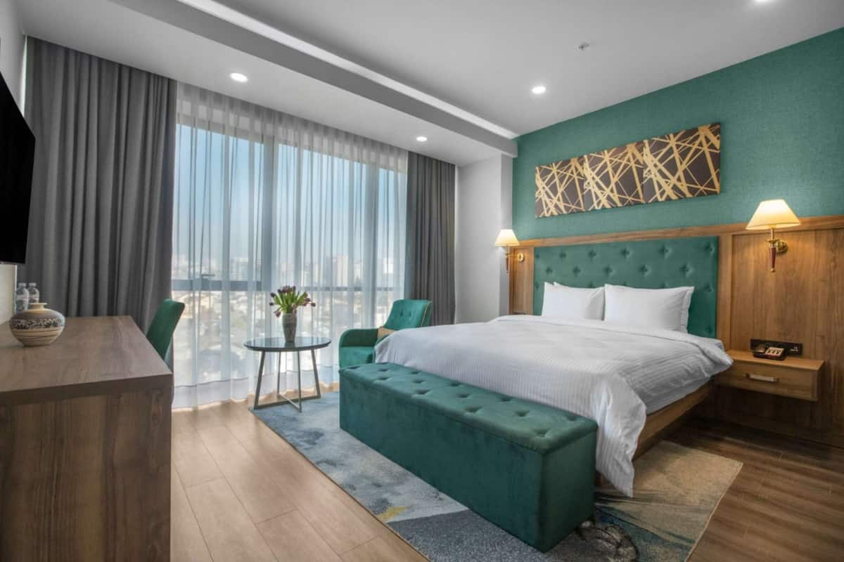 A modern hotel room with a large bed, a green upholstered bench, a small table with a vase, and a view of the city through large windows.