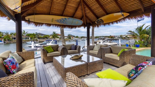 An outdoor lounge area with wicker furniture, a glass-top table, and surfboards hanging from the ceiling. - Paradise on Banksia Canal (BANKSIA BEACH)