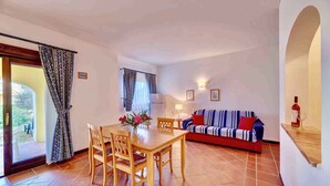 A dining area with a wooden table and chairs, a sofa with a striped blanket, and a bottle on a shelf. - Holiday home mit Klimaanlage (Stintino)