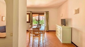 A dining area with a table and chairs, a cabinet with a television, and a view of buildings through a window. - Holiday home mit Klimaanlage (Stintino)