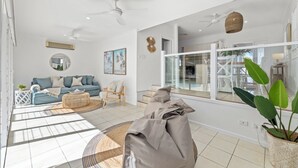 A spacious living room with a blue sofa, a round wicker coffee table, and a grey pouf. There are two ceiling fans, a large window with a view of the outside, and a potted plant. - Fairways Beach House - Stylish Beach House, Pet Friendly, Pool, Walk to Cafes, Beach (Sawtell)