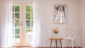A room with a wooden floor, white curtains, a white chair, a small round table with a vase and a pitcher, and a framed picture on the wall. - Fairways Beach House - Stylish Beach House, Pet Friendly, Pool, Walk to Cafes, Beach (Sawtell)