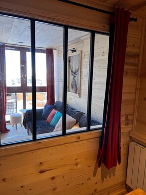 L’intérieur d’un chalet en bois, avec une grande fenêtre, un canapé noir agrémenté de coussins colorés, un petit tabouret et un tableau représentant un cerf accroché au mur.