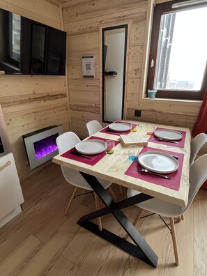 Une pièce aux murs lambrissés, avec une table à manger dressée pour quatre personnes, des chaises blanches, un téléviseur à écran plat fixé au mur et une fenêtre donnant sur des immeubles.