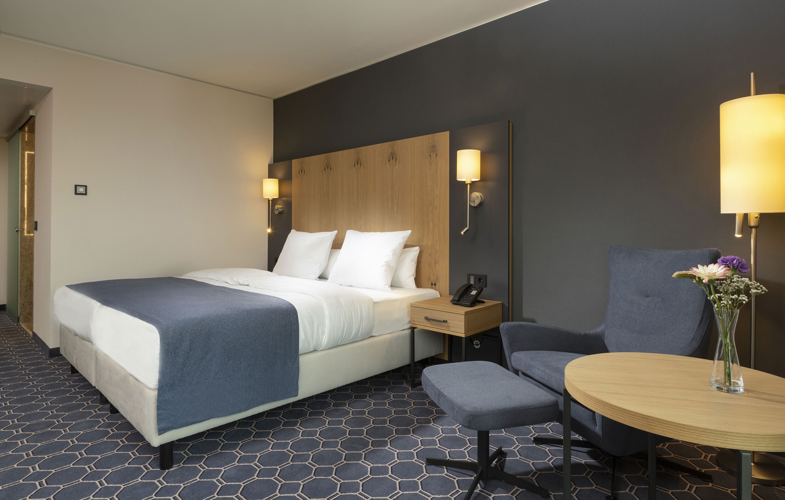 A hotel room with a large bed, bedside table with a phone, a small round table with a vase, and a chair.