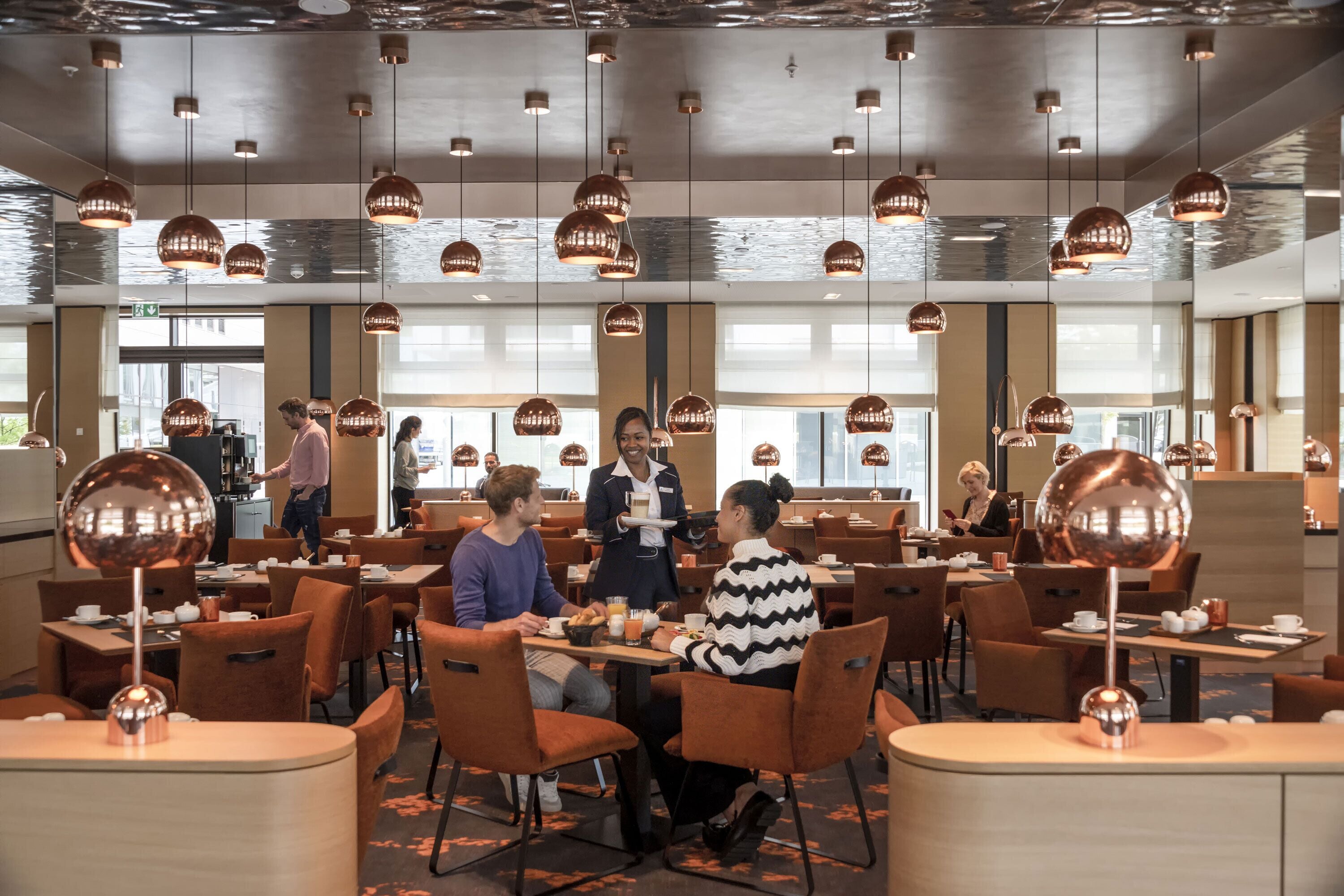 A modern dining area with multiple tables and chairs, overhead lighting, and a view of the city through large windows.