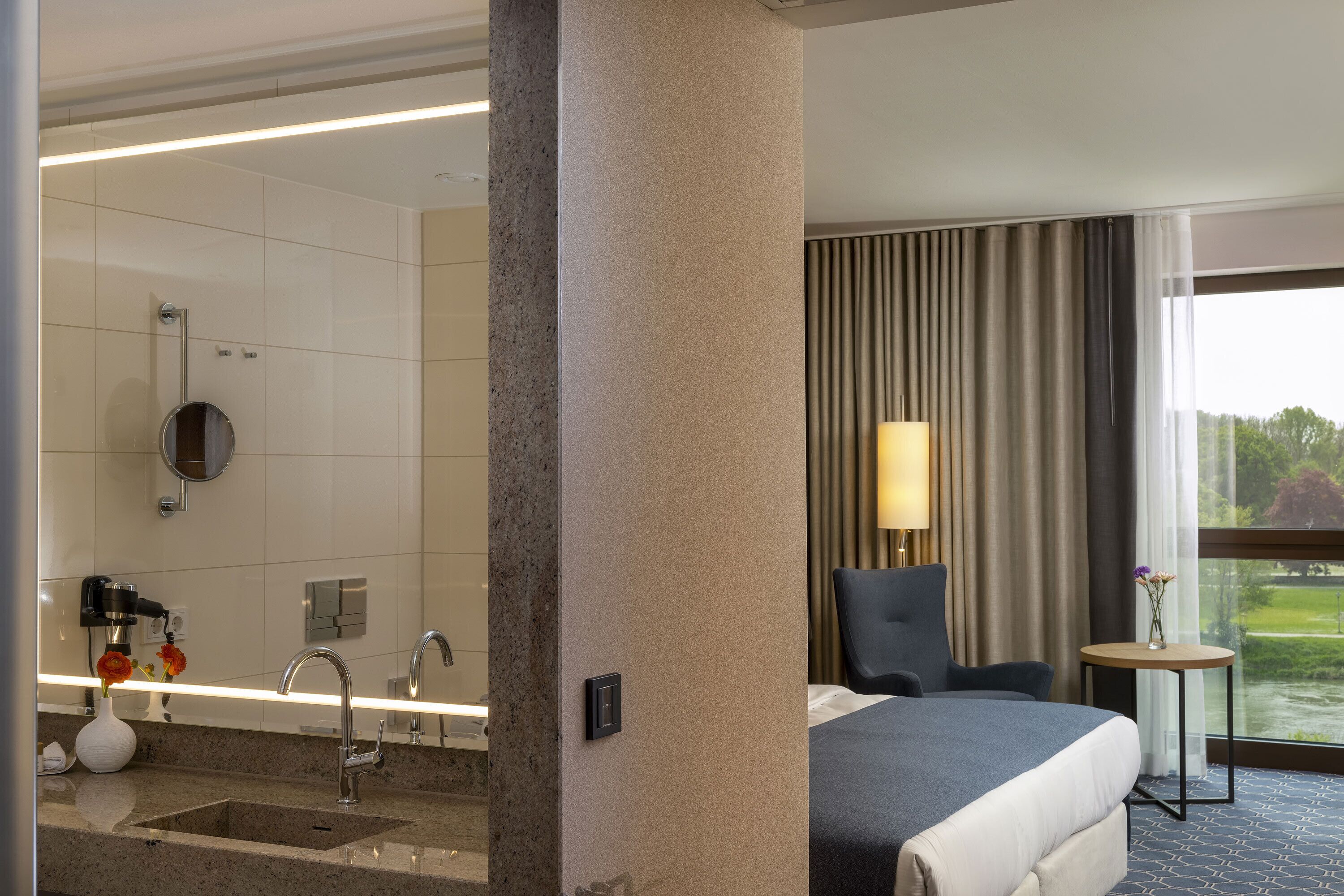 Ein Hotelzimmer mit Bett, einem kleinen Tisch mit Lampe, einem Badezimmer mit Waschbecken und Spiegel sowie Blick auf Grünfläche.