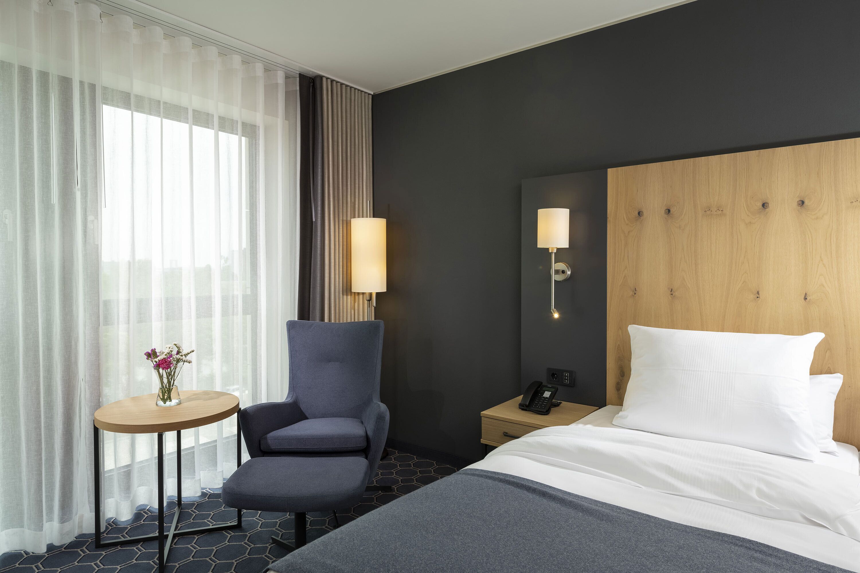 A hotel room with a bed, a chair, a small table with a vase of flowers, a nightstand with a lamp, and a view of the city through sheer curtains.