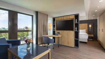 Quarto de hotel moderno com uma janela grande, uma secretária com cadeira, uma cama e uma televisão fixada na parede.