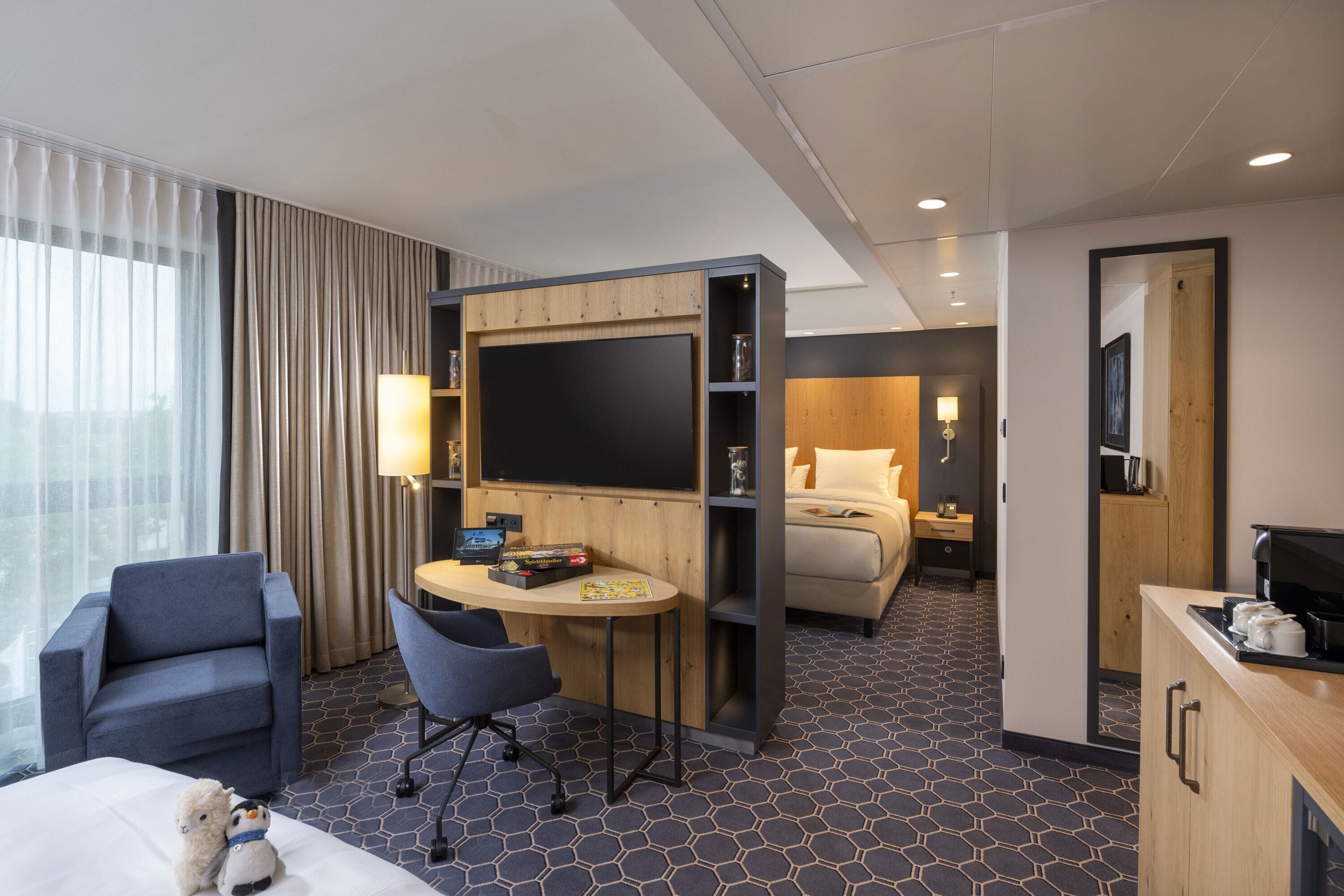 A hotel room with a bed, a desk with a computer, a chair, a small table, and a TV.