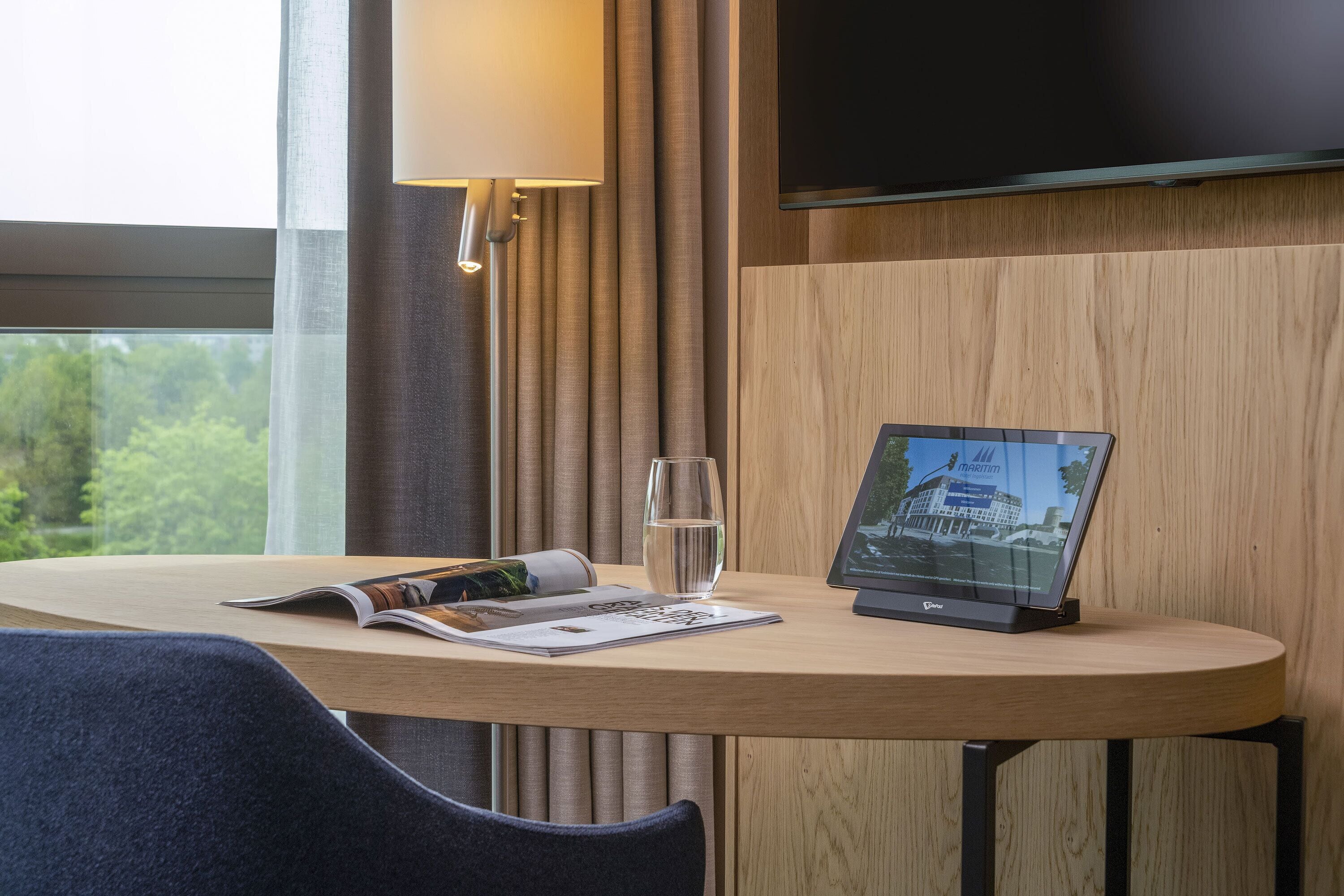 A hotel room with a wooden table, a magazine, a glass of water, a laptop, and a TV mounted on the wall.