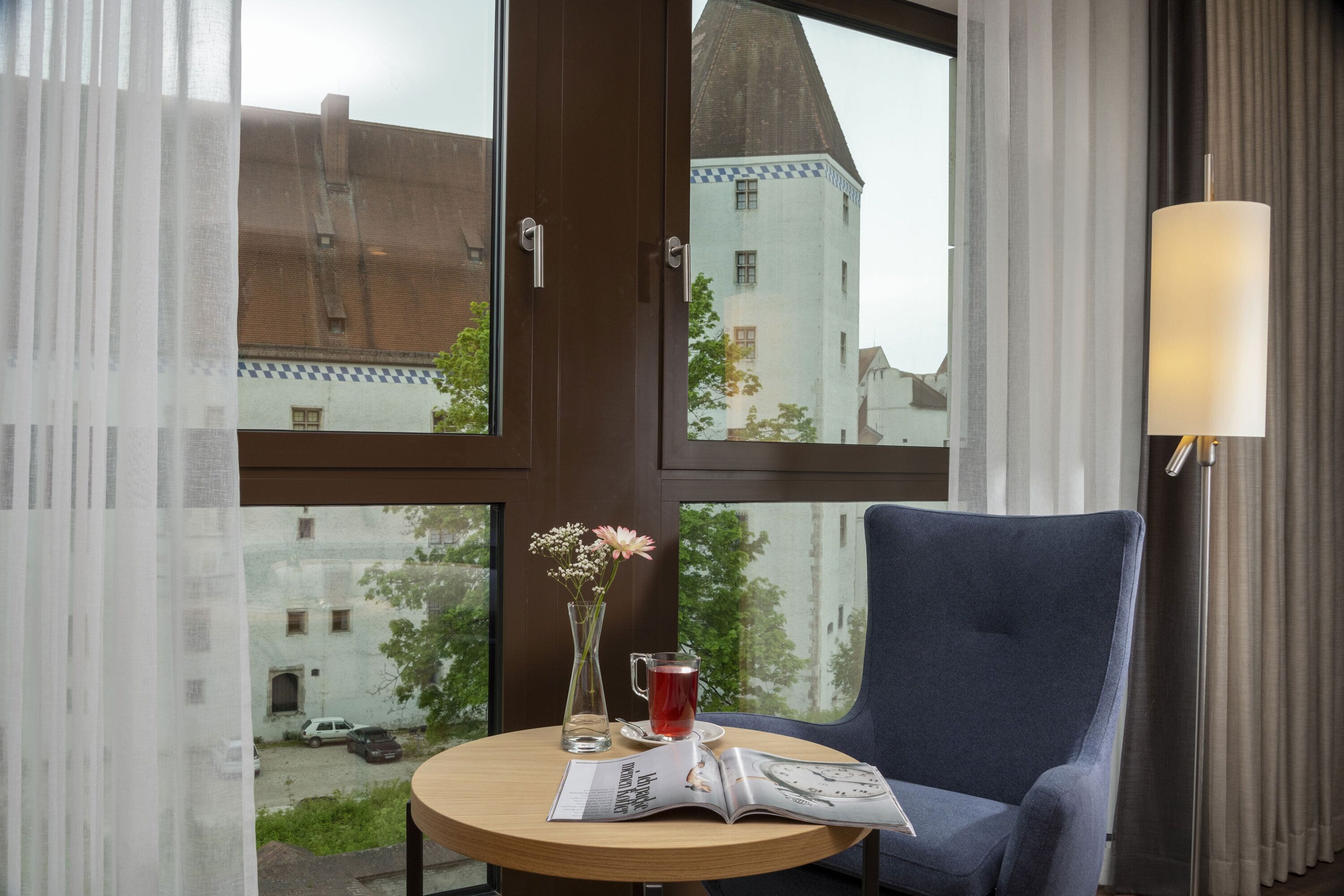 A hotel room with a round table, a blue armchair, a vase with flowers, a glass of red drink, and a magazine.