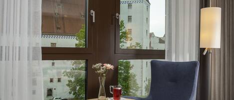Habitación de hotel con una mesa redonda, un sillón azul, un florero con flores, un vaso de bebida roja y una revista.