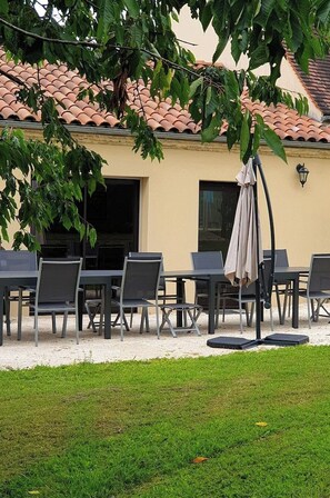 An outdoor seating area with tables and chairs, a covered umbrella, and a building with a tiled roof in the background.