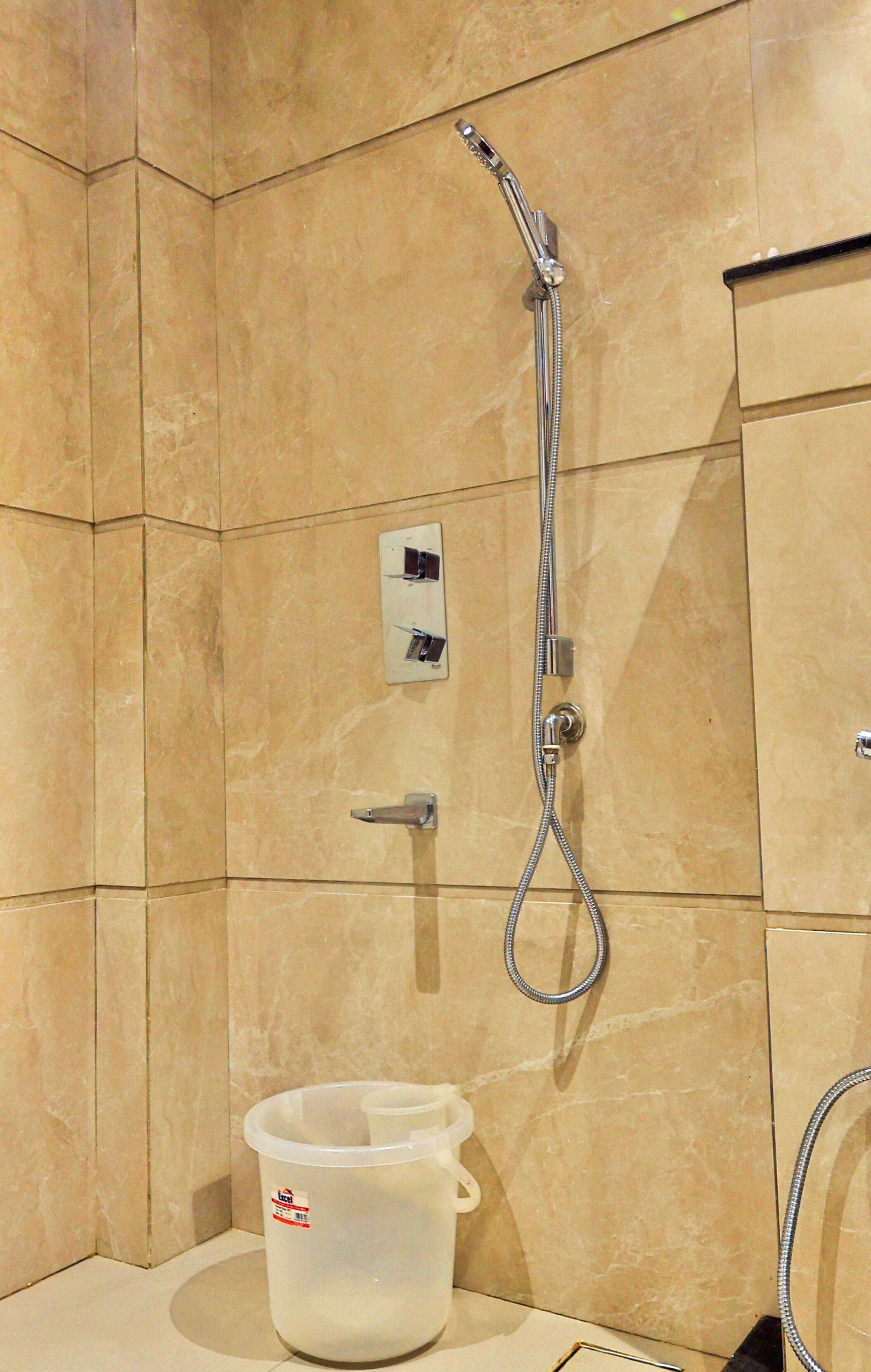 A bathroom with beige tiled walls, a shower with a handheld hose, a soap dispenser, and a plastic bucket on the floor.