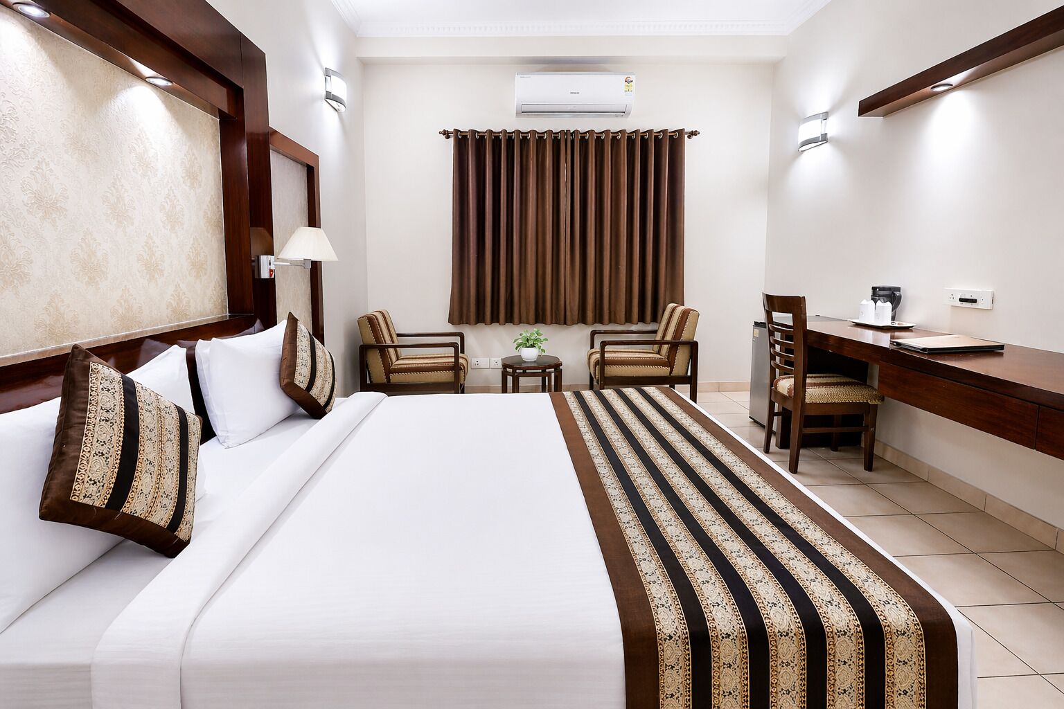 A hotel room with a bed, two chairs, a desk, a small table, and an air conditioning unit.